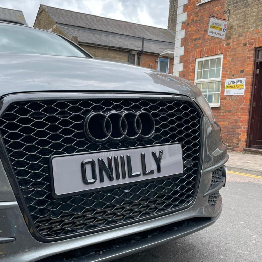 4D retro tinted show plate featuring raised gloss black lettering spelling ONIILLY, fitted to a grey Audi with a black honeycomb grille outside Bedford Number Plates.